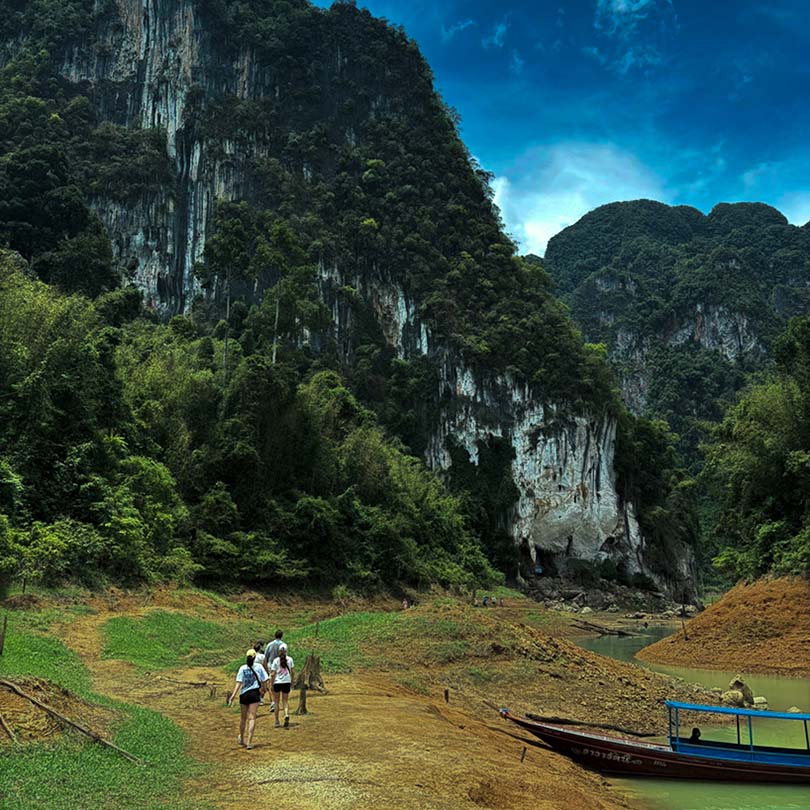 Group of hikers walking along a muddy trail toward towering limestone cliffs and dense greenery, a boat docked on the river edge.