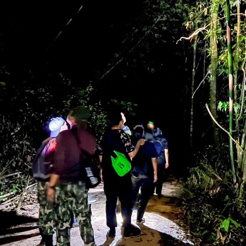 A group of hikers walking along a muddy forest trail at night, carrying backpacks under sparse light.